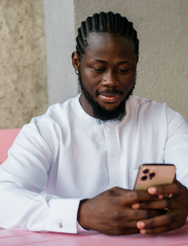 ein Schwarzer junger Mann mit Cornrows, Bart, und in einem weissen Hemd, schaut entspannt in sein Smartphone