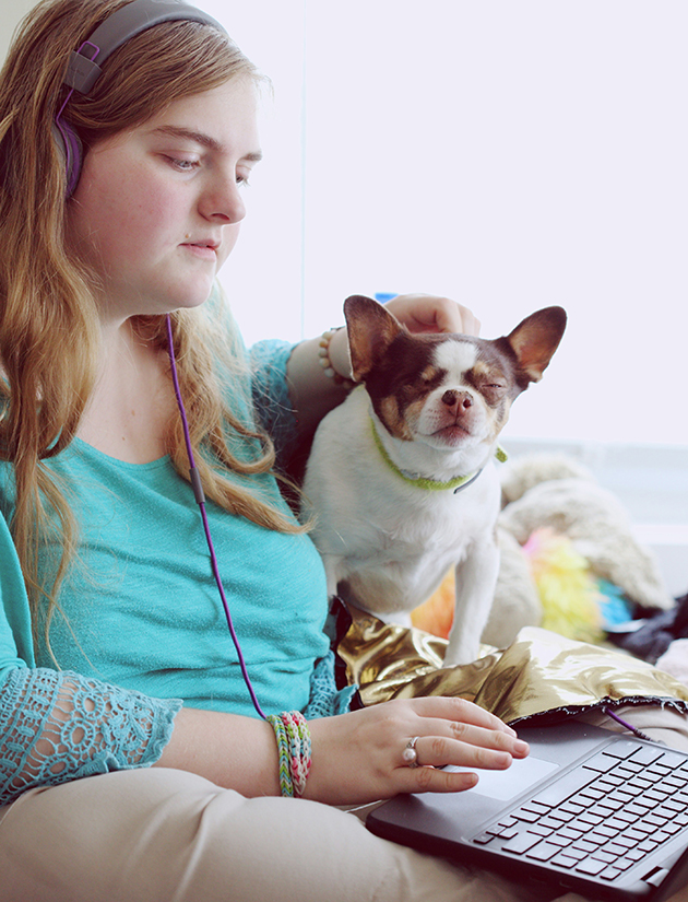 Ein junges Mädchen sitzt auf der Couch neben ihrem Hund, den sie krault. Sie hört Musik über große Kopfhörer, die mit ihrem Laptop verbunden sind, den sie auf ihrem Schoß hat.