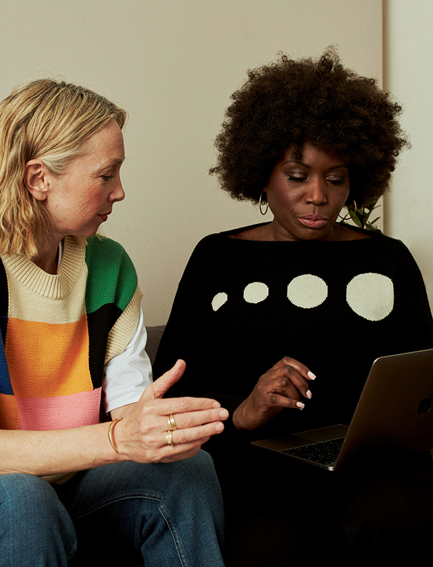 Eine Szene aus dem Arbeitsalltag einer Beratungsstelle zwischen zwei Fachleuten: Zwei Frauen mittleren Alters sitzen nebeneinander und besprechen ein Thema. Die Schwarze Frau hat einen Laptop auf dem Schoß, die weisse Frau schaut auf den Bildschirm. Beide sind in das Thema vertieft und in führen ein fachliches Gespräch.