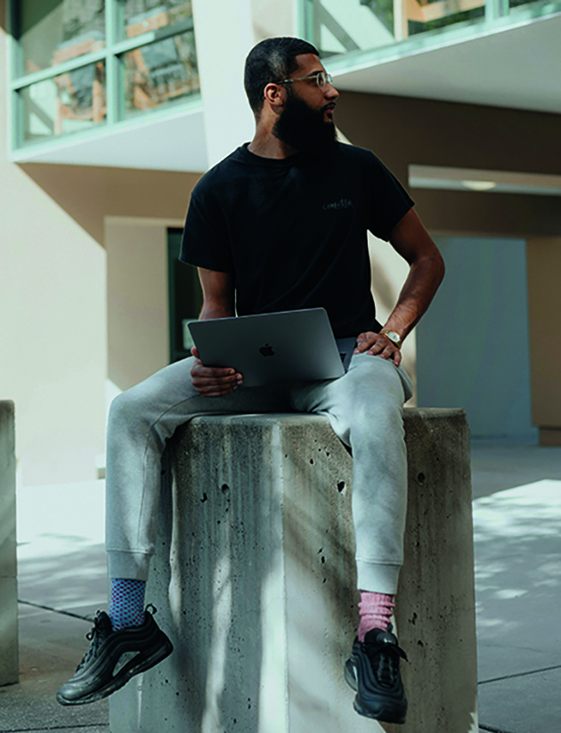 Junger Mann mit Bart und Brille, locker gekleidet mit Jogginghose und zwei verschiedenfarbigen Socken, sitzt auf einem Betonblock, seinen Laptop auf dem Schoß in einem städtischen Umfeld und schaut an der Kamera vorbei zu jemanden hin.