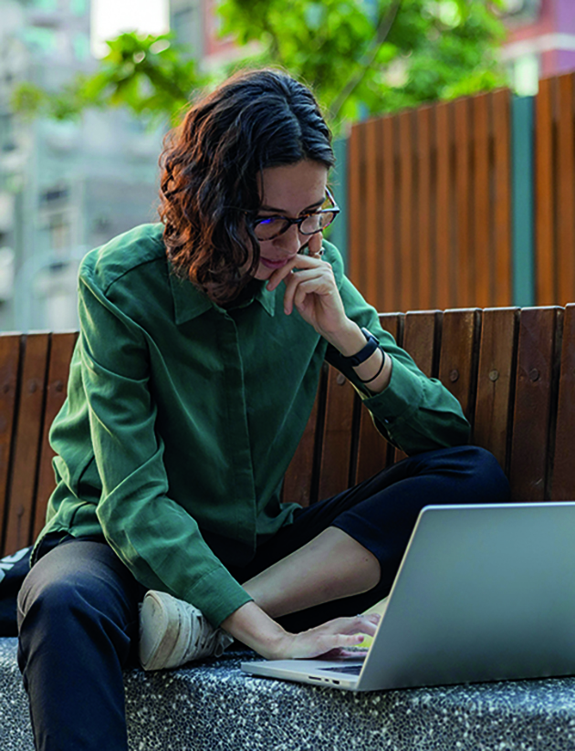 Eine junge Frau in einem grünen, lockeren Hemd, Sneakern und Brille, sitzt auf einer Parkbank –sie schaut konzentriert in ihren Laptop, der vor ihr steht